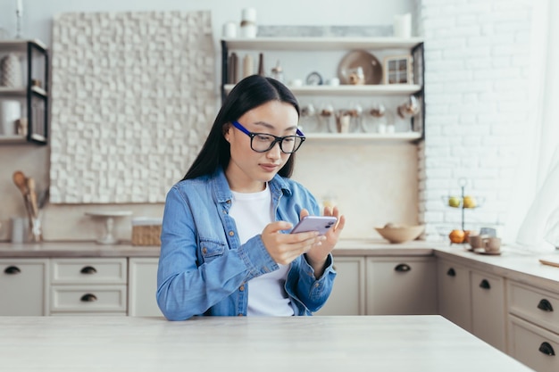 Joven hermosa mujer asiática sentada en la cocina en casa y usando el teléfono móvil se desplaza por el