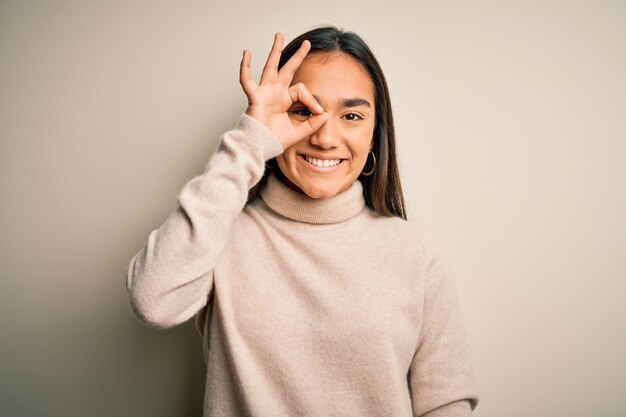 Joven hermosa mujer asiática con suéter casual de cuello alto sobre fondo blanco haciendo un buen gesto con la mano sonriendo mirando a través de los dedos con cara feliz