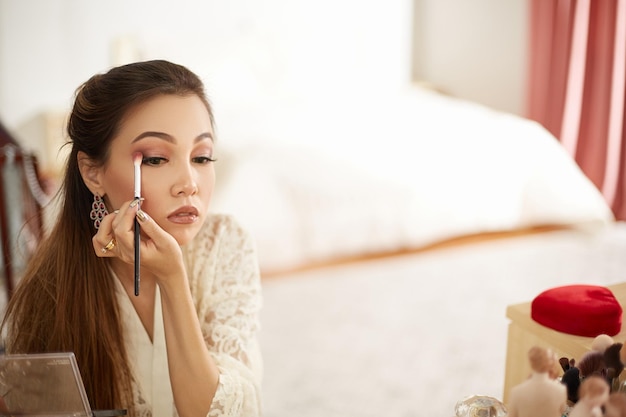 Una joven y hermosa mujer asiática con un vestido de encaje beige se maquilla por la mañana y usa rubor de sombra de ojos y Fundación en el dormitorio profundidad superficial de enfoque enfoque selectivo