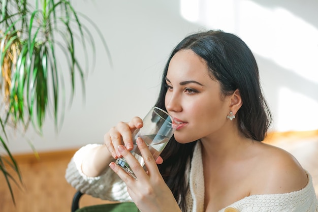 Una joven y hermosa mujer de cabello oscuro con un cárdigan de punto bebe agua limpia de un vidrio Retrato de modelo de casa acogedora Salud