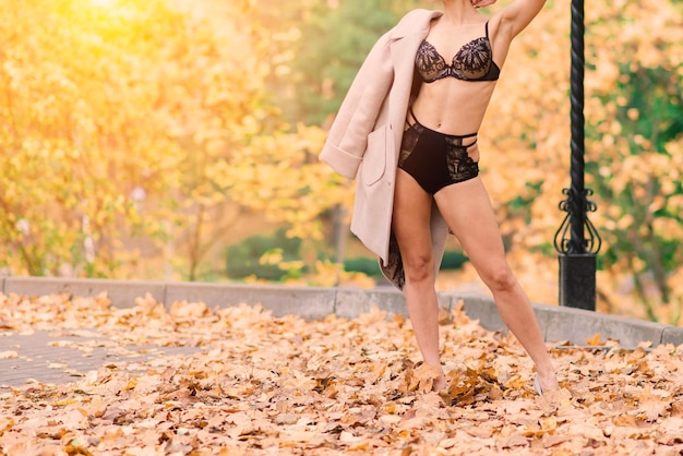 Joven hermosa mujer de cabello oscuro con figura esbelta en lencería y abrigo clásico en el parque de otoño