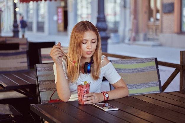 Joven hermosa mujer caucásica de unos 25 años con cabello rubio está sentada al aire libre y comiendo postre de frutas mientras mira la pantalla del teléfono móvil.