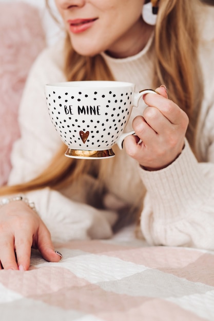 Joven hermosa mujer caucásica con cabello largo castaño en suéter blanco sostiene una taza blanca de café o té, lo bebe y se acuesta en el sofá