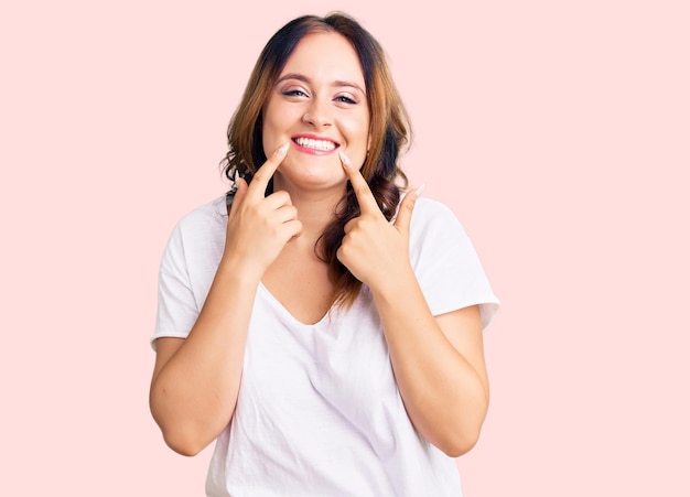 Joven hermosa mujer caucásica con camiseta blanca casual sonriendo con los dedos de la boca abierta señalando y forzando una sonrisa alegre