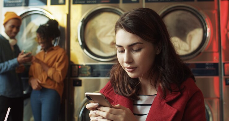 Foto joven hermosa mujer caucásica golpeando y enviando mensajes de texto en el teléfono inteligente mientras está sentado en la sala de servicio de lavandería. elegante mujer bonita escribiendo en el teléfono y esperando ropa para lavar en la máquina.