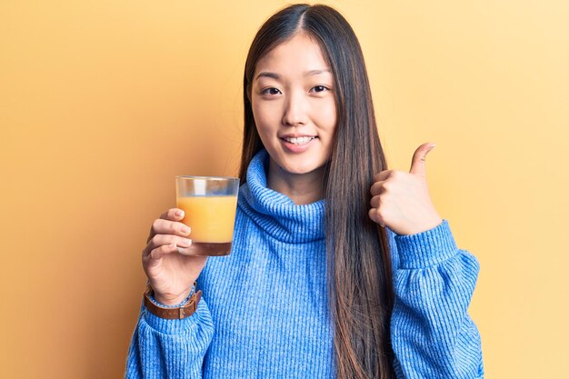 Joven hermosa mujer china bebiendo un vaso de jugo de naranja sonriendo feliz y positiva, pulgar arriba haciendo excelente y signo de aprobación