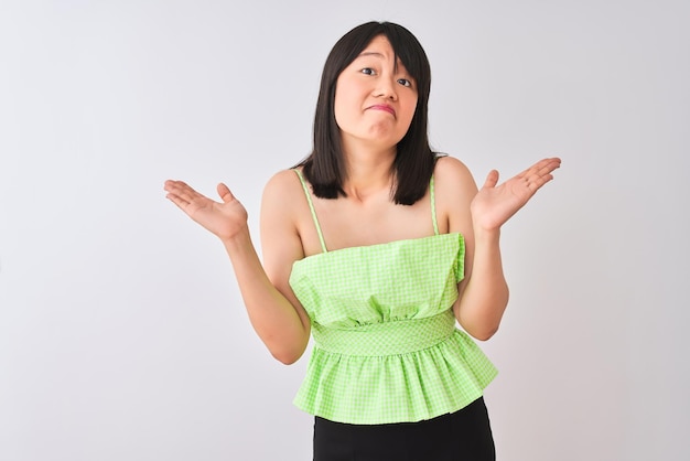 Joven hermosa mujer china con camiseta verde sobre fondo blanco aislado expresión desorientada y confusa con los brazos y las manos levantadas Concepto de duda