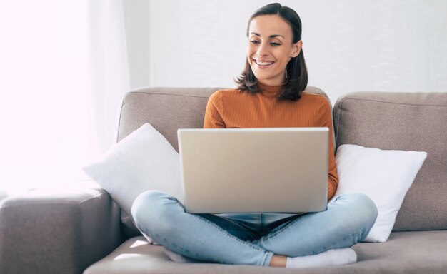 Joven hermosa mujer confiada que trabaja con su computadora portátil mientras está sentado en el sofá en casa