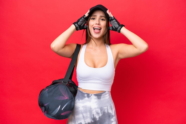 Joven hermosa mujer deportiva con bolsa deportiva aislada de fondo rojo con expresión de sorpresa