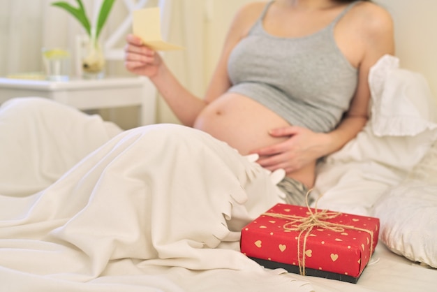 Joven hermosa mujer embarazada con caja de regalo, mujer sentada en su casa en la cama leyendo una tarjeta de felicitación