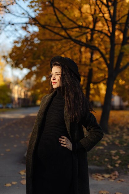 Joven hermosa mujer embarazada con un sombrero con el pelo oscuro en un vestido ajustado negro y un abrigo