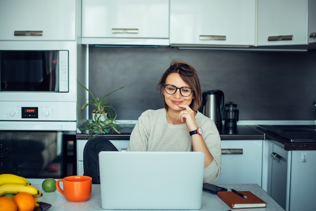 Joven hermosa mujer europea con gafas elegantes trabaja con un portátil en la cocina de casa. Comuníquese con amigos y familiares en línea a través de Internet. Interior minimalista en tonos beige.