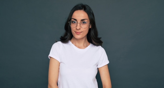 Joven hermosa mujer feliz con estilo en una camiseta blanca y anteojos está planteando sobre el fondo gris