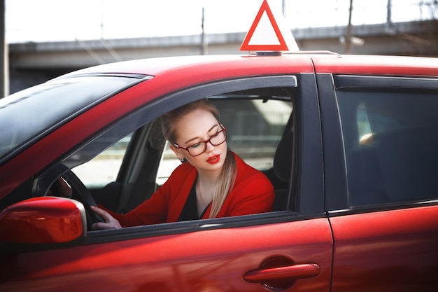 Joven hermosa mujer feliz en rojo sentado al volante nuevo coche de entrenamiento.