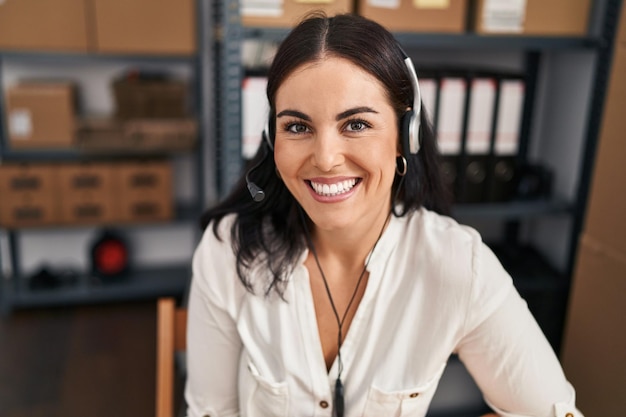 Joven hermosa mujer hispana agente de comercio electrónico sonriendo confiada sentada en la mesa en la oficina