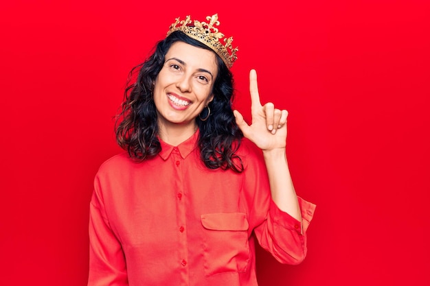 Joven hermosa mujer hispana con corona de princesa sonriendo con una idea o pregunta señalando con el dedo hacia arriba con la cara feliz número uno