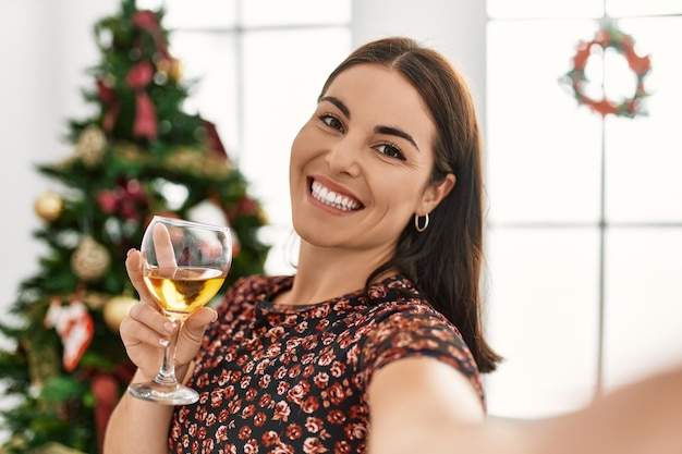 Joven hermosa mujer hispana hace selfie con cámara de pie junto al árbol de Navidad en casa