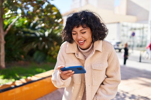 Joven hermosa mujer hispana sonriendo confiada viendo videos en un teléfono inteligente en el parque