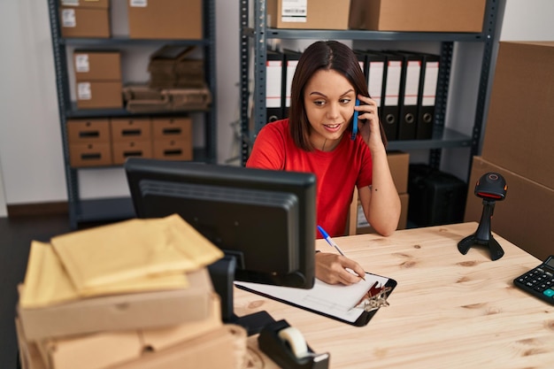 Foto joven hermosa mujer hispana trabajadora de comercio electrónico hablando en un teléfono inteligente escribiendo en un documento en la oficina