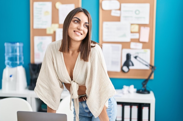 Joven y hermosa mujer hispana trabajadora de negocios sonriendo segura de pie junto al escritorio en la oficina