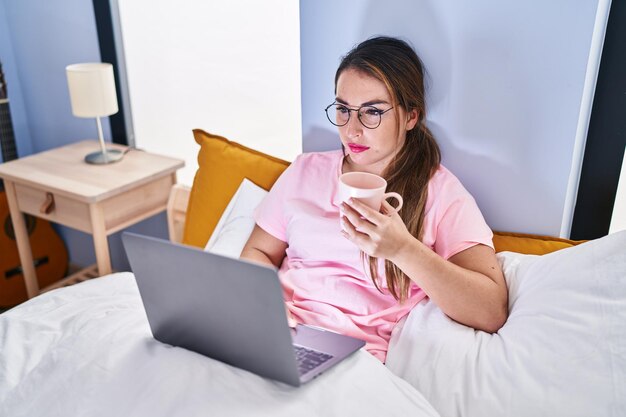 Joven hermosa mujer hispana usando laptop tomando café en el dormitorio