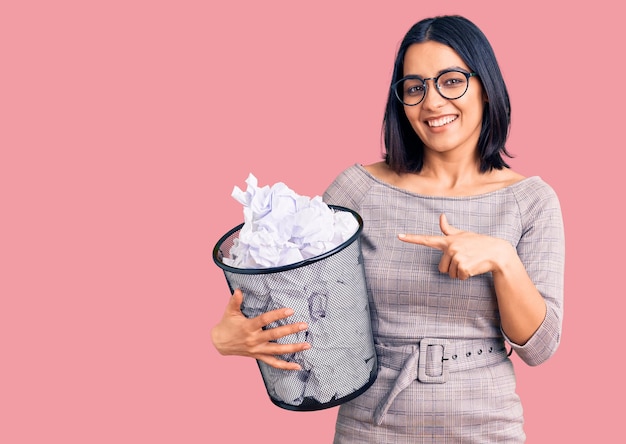 Joven hermosa mujer latina sosteniendo papelera llena de papeles arrugados sonriendo feliz señalando con la mano y el dedo