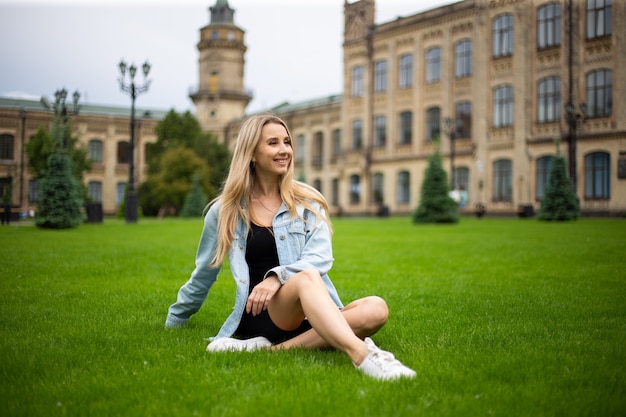 Joven hermosa mujer de moda moderna en una chaqueta de mezclilla se sienta en un césped verde