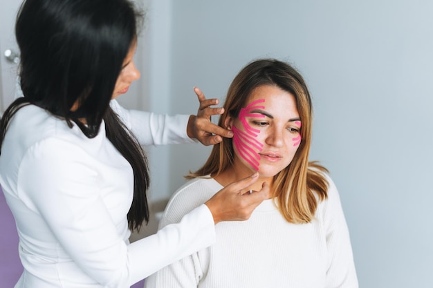 Joven hermosa mujer morena con el médico cosmetólogo de cabello largo hace cinta facial para el paciente en el consultorio Las mujeres jóvenes realizan procedimientos antienvejecimiento con la ayuda de la cinta kinesio