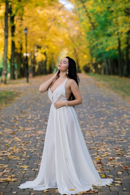 Joven hermosa mujer morena con un vestido blanco largo en el parque de otoño.