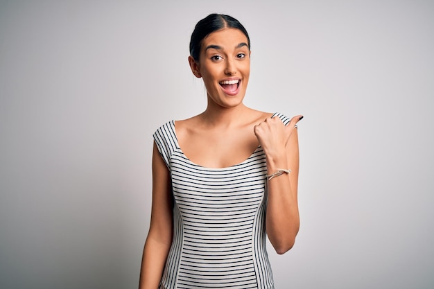 Joven hermosa mujer morena con un vestido informal a rayas sobre un fondo blanco aislado sonriendo con cara feliz mirando y apuntando hacia un lado con el pulgar hacia arriba