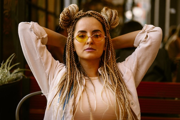 Joven hermosa mujer multirracial con peinado afro rubio trenzas zizi en el café de la calle. gafas de sol amarillas, maquillaje brillante, estilo hippie