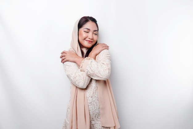 Joven hermosa mujer musulmana asiática con un pañuelo en la cabeza sobre fondo blanco abrazándose a sí misma feliz y positiva sonriendo confiada Selflove y selfcare