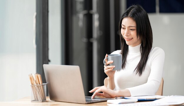 Joven hermosa mujer de negocios asiática sonriente sosteniendo una taza de café y usando una mesa portátil en la oficina