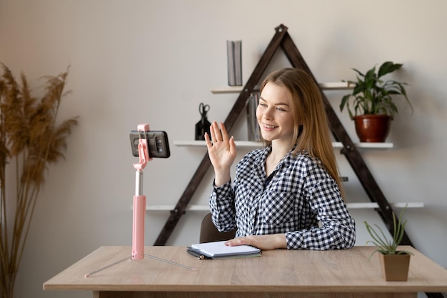 Una joven y hermosa mujer de negocios caucásica está sentada en la mesa para comunicarse por videoconferencia levantó la mano con un gesto de trípode de saludo para teléfono inteligente Videollamada en casa desde la oficina