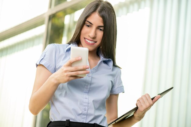 Joven hermosa mujer de negocios sonriente con teléfonos inteligentes y planificador de pie frente al edificio de oficinas