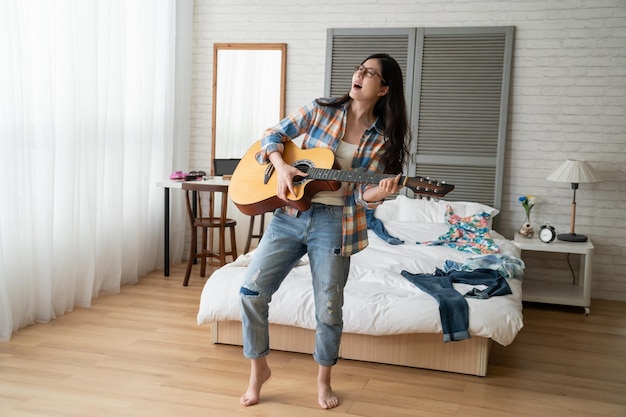 Foto joven hermosa mujer oriental de pie en el suelo de madera tocando la guitarra cantando disfrutar de la música escrita por ella misma. girl power divirtiéndose concepto estilo de vida quedarse en casa tiempo libre. linda mujer divirtiéndose