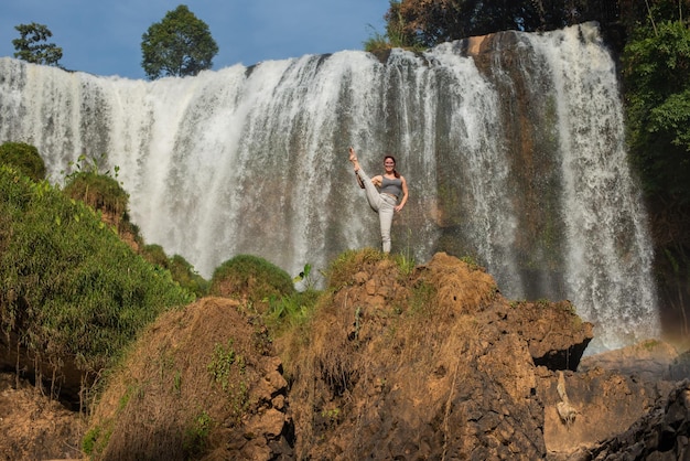 Una joven y hermosa mujer pelirroja con anteojos se para en una cuerda en un acantilado cerca de una cascada La niña practica yoga en la naturaleza Pilates en la naturaleza salvaje