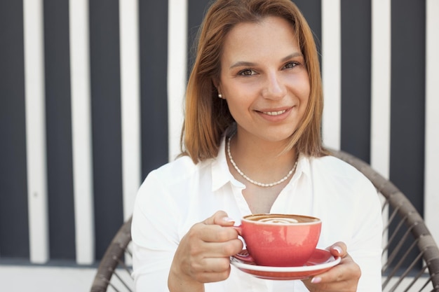 Joven hermosa mujer pelirroja con camisa blanca relajándose en un café de la calle después del trabajo y bebiendo café