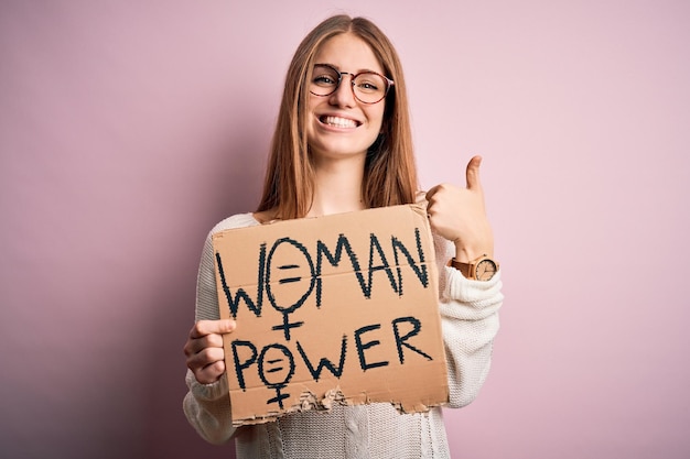 Joven hermosa mujer pelirroja pidiendo derechos de las mujeres sosteniendo una pancarta sobre fondo rosa feliz con una gran sonrisa haciendo bien firmar el pulgar hacia arriba con los dedos excelente señal