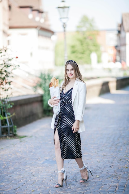 Joven hermosa mujer de pelo largo de 25 años con un vestido blanco y negro compra una baguette francesa en el centro de una ciudad europea Panadería fresca Modelo elegante camina por la ciudad