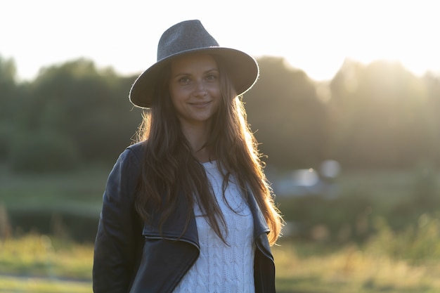 Una joven y hermosa mujer de pelo largo con un sombrero sonríe, mira a la cámara en el parque en el brillante resplandor de los rayos del atardecer