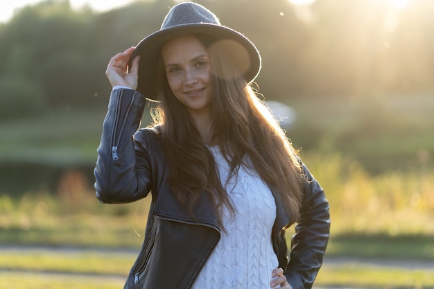 Una joven y hermosa mujer de pelo largo con un sombrero sonríe, mira a la cámara en el parque en el brillante resplandor de los rayos del atardecer