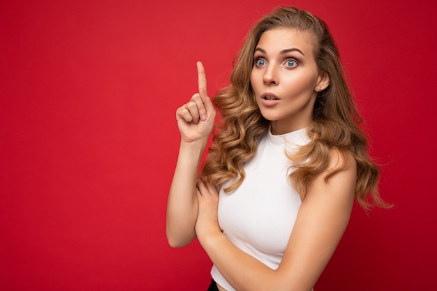 Joven hermosa mujer rizada rubia positiva sorprendida con emociones sinceras vistiendo top blanco aislado sobre fondo rojo con espacio libre y apuntando al espacio de la copia para el texto.