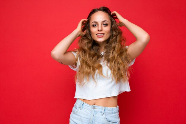 Joven hermosa mujer rubia con camiseta blanca mirando a la cámara. La mujer positiva muestra emociones faciales sinceras. Modelo divertido aislado en la pared roja con espacio vacío.