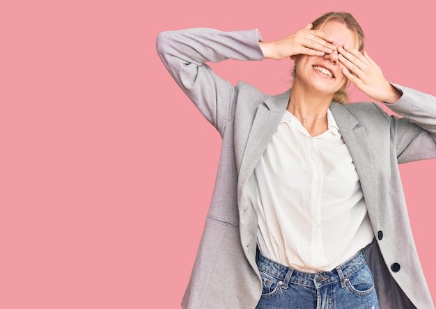 Joven hermosa mujer rubia con chaqueta elegante que cubre los ojos con las manos sonriendo alegre y divertida. concepto ciego.