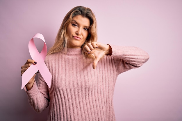 Joven hermosa mujer rubia pidiendo apoyo para el cáncer sosteniendo el símbolo de la cinta rosa con el signo negativo de la cara enojada que muestra disgusto con el concepto de rechazo de los pulgares hacia abajo