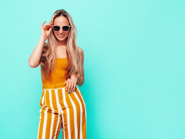 Joven hermosa mujer rubia sonriente en ropa amarilla de moda de verano Mujer despreocupada sexy posando junto a la pared azul en el estudio Modelo positivo divirtiéndose en el interior Alegre y feliz En gafas de sol