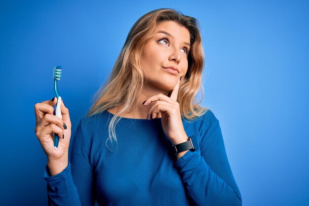 Joven hermosa mujer rubia usando cepillo de dientes sobre fondo azul aislado cara seria pensando en la pregunta idea muy confusa