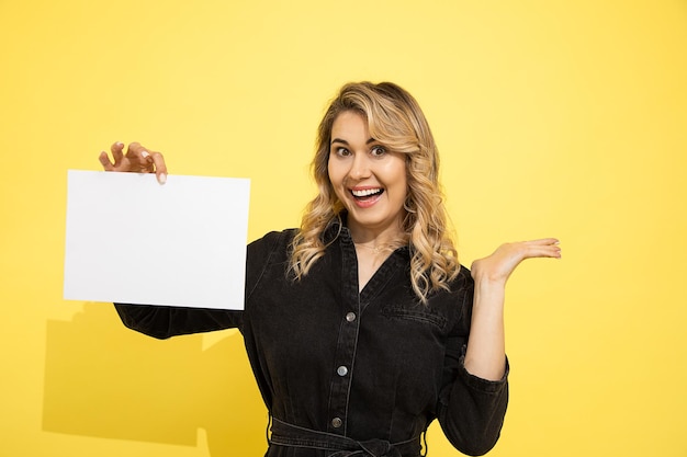 Joven hermosa mujer rubia vestida de negro tiene hoja vacía para inscripción sobre fondo amarillo en manos apuntando con la mano, sonríe y ríe felizmente sobre fondo amarillo en estudio