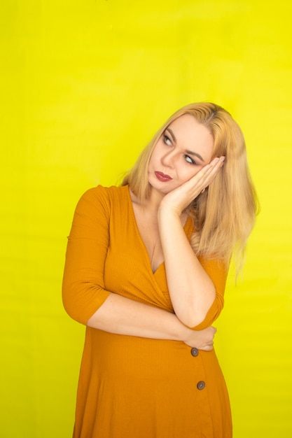 Joven hermosa mujer rubia en vestido amarillo oscuro posando sobre pared amarilla interior